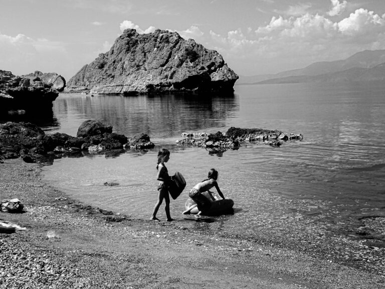 Δύο παιδιά κουβαλούν πράγματα σε βραχώδη παραλία, με μια μεγάλη βραχονησίδα πίσω τους.