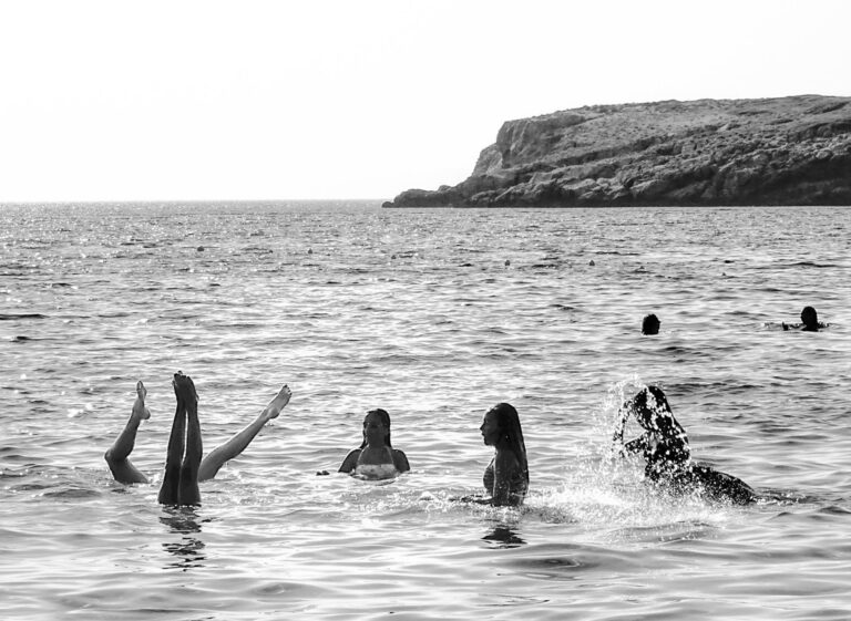 Λουόμενοι στη θάλασσα, ένας κάνει κατακόρυφο με τα πόδια στον αέρα, με γκρεμούς στον ορίζοντα.