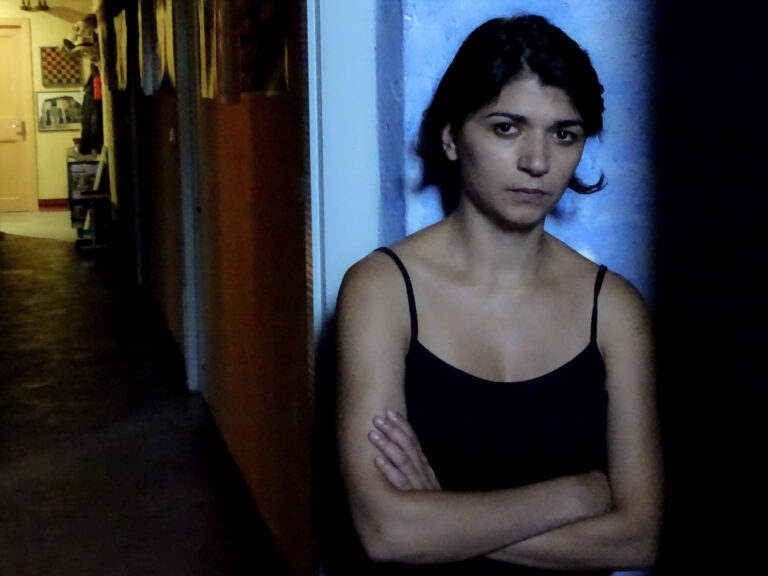 Γυναίκα με μαύρο μπλουζάκι και σταυρωμένα χέρια, σε μπλε φως στο τέλος ενός διαδρόμου.