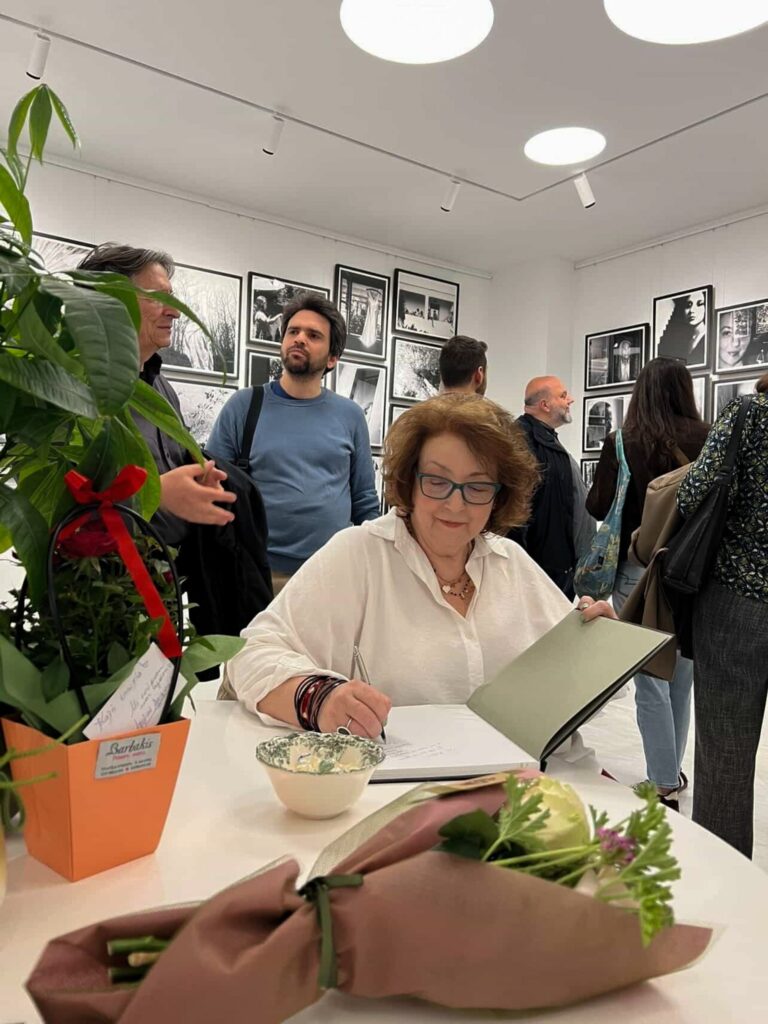 Γυναίκα με γυαλιά υπογράφει βιβλία σε τραπέζι με λουλούδια, με καλεσμένους όρθιους πίσω της.