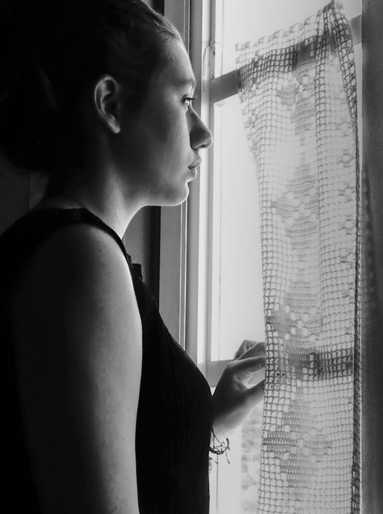 Γυναίκα με σκούρο μπλουζάκι κοιτάζει έξω από παράθυρο με πλεκτή κουρτίνα.