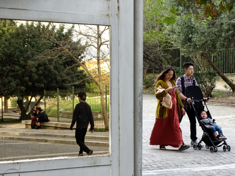 Οικογένεια με καρότσι περπατά δίπλα σε ένα παράθυρο που καδράρει ένα μονοπάτι πάρκου.