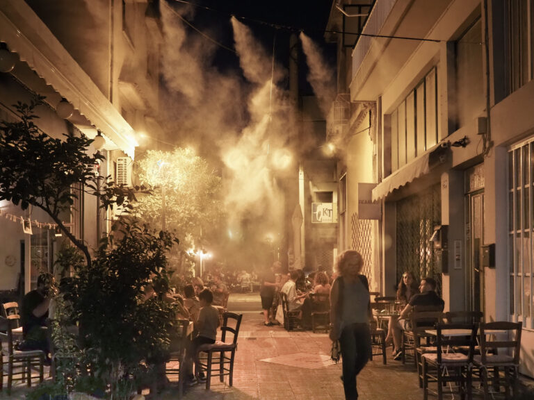 Στενός δρόμος με τραπεζάκια καφέ τη νύχτα, με σύννεφο από ψεκασμό νερού να λάμπει στα φώτα.
