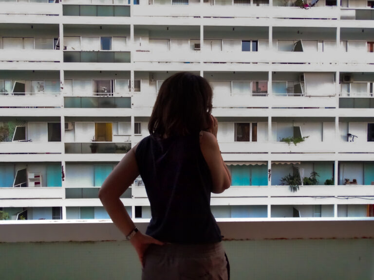 Γυναίκα σε μπαλκόνι από την πλάτη, απέναντι από μια μεγάλη πολυκατοικία.
