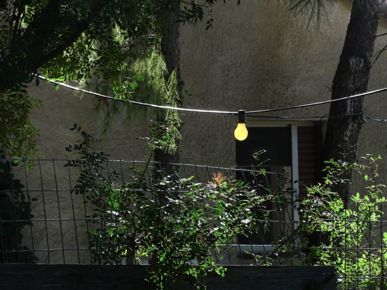 Κήπος το σούρουπο, με σειρά από αναμμένα λαμπάκια πάνω από φράχτη και πράσινους θάμνους.