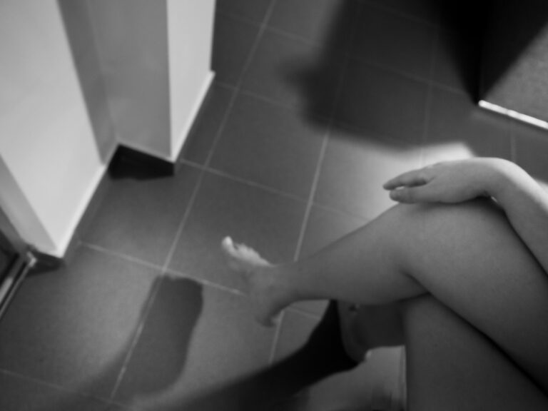 Γυμνό: γυναίκα καθισμένη σε πλακάκια, με το χέρι στο γόνατο, δίπλα σε μια πόρτα.